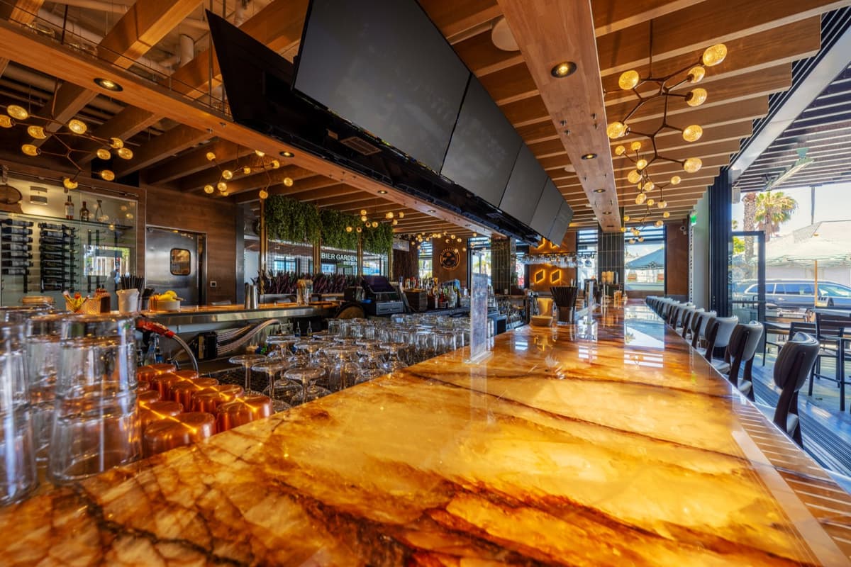 The backlit onyx bar with rows of glassware and overhead screens at Bier Garden Oceanside